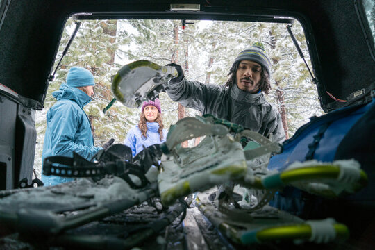Young African-American male lifts his snowy skis out of the trunk while his friends talk