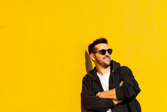 Young bearded man standing against a yellow wall with crossed arms