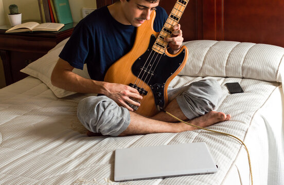 Casual man sitting on a bed playing electric bass at home.
