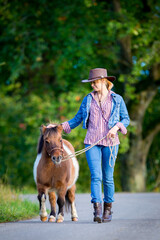 Girl in Western clothing is walking with a horse outdoor. Young woman leads a Shetland pony along the road in summertime.
