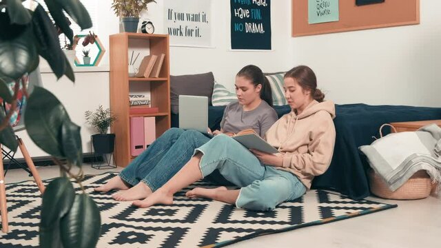Tracking shot of young female college students sitting together on floor by bed in their cozy dorm and doing assignment