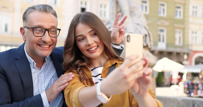 Pretty young Caucasian girl taking selfie photos on smartphone camera with father at street in town. Middle-aged handsome man in glasses smiling to mobile phone as making photo with adult daughter.