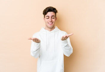 Young caucasian man isolated on beige background holding something with palms, offering to camera.