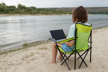 young female with laptop in her knees is sitting camping chair on the bank of picturesque river. Frelancer and remote work concept. Opportunity for distant work.