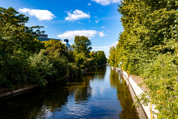 Fototapeta na wymiar A sunny day in Berlin, Germany. Image taken from a bridge in the city center features one of many canals inside the city with trees on both sides, reflection in water and clear sky in background