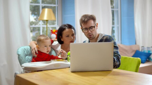 Happy young couple feeding baby boy in highchair and using laptop at home