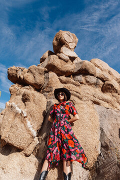 Young stylish cowgirl in dress leans against rock pile in desert.