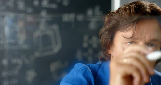 Close up Caucasian man mathematician writing formulas and laws on transparent screen in classroom. Blackboard with digits and drawing on background. Male teacher or scientist counting and explaining.