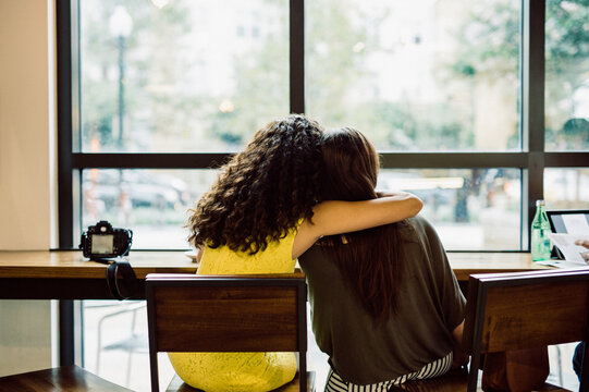 Two women embrace while sitting at a cafe and looking out the window