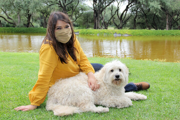 
latin woman with protection mask sitting on the grass with white dog, walking on the shore of the lake, new normal