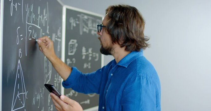 Close up of Caucasian male teacher in glasses writing formulas and math laws on blackboard and looking at smartphone. Educative mathematics lection. Man lecturer using mobile phone as cribe.