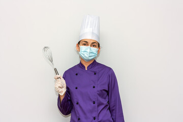 Naklejka premium Woman chef with surgical mask and gloves for coronavirus prevention looks at the camera and holds a whisk in her hand.