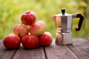 Geyser coffee maker stands on a wooden board against the green blurred background. With red apples