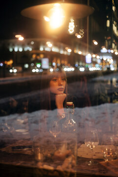 Blurred night portrait of young beautiful woman sitting alone in cafe