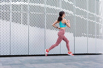 Outdoor summer street portraits of young beautiful sexy sport style brunette woman exercising, wearing blue and red outfit. Wearing wireless earphones. Modern metal architecture. Lifestyle sports, running, cardio training.