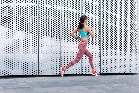 Outdoor summer street portraits of young beautiful sexy sport style brunette woman exercising, wearing blue and red outfit. Wearing wireless earphones. Modern metal architecture. Lifestyle sports, running, cardio training.