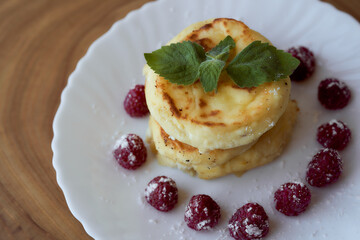 Juicy curd cheesecakes lie on a white saucer with raspberries and mint leaf. Light background