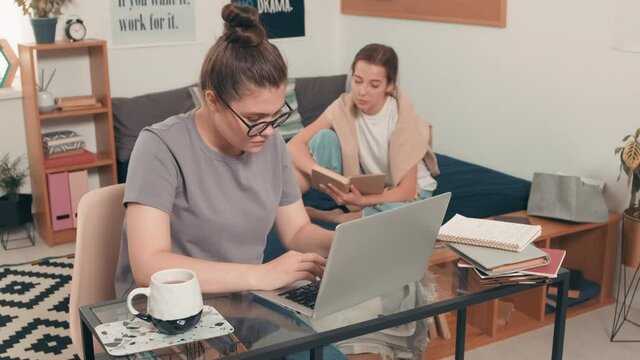 Tracking shot of young female student in glasses sitting at desk in dormitory and typing on laptop. Her female roommate sitting on bed and reading textbook