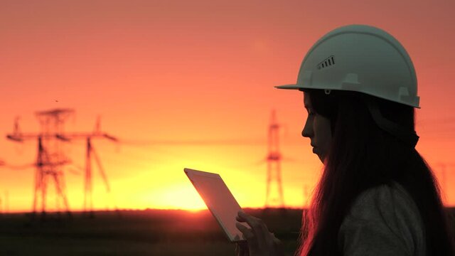 A woman power engineer in white helmet inspects power line using data from electrical sensors on tablet. High voltage electrical lines at sunset. Distribution and supply of electricity. clean energy