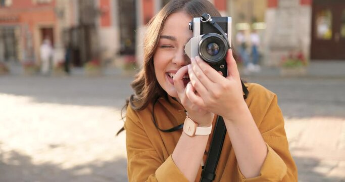 Close up of pretty young Caucasian girl taking photo with camera at street of town. Outdoor. Female cheerful photographer outside in city on sunny summer day.