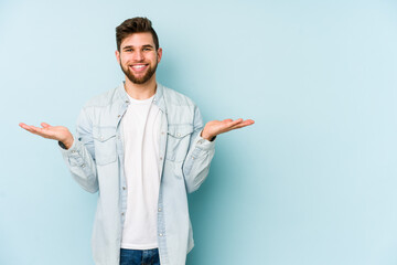 Young caucasian man isolated on blue background makes scale with arms, feels happy and confident.