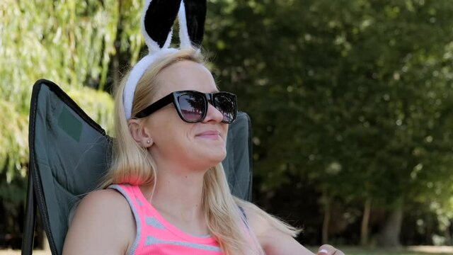 Young woman in sunglasses with funny rabbit ears hat sits in a chair and rests. Close-up. Outdoors.