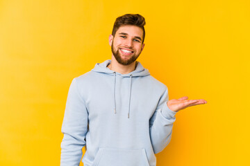 Young caucasian man isolated on yellow background showing a copy space on a palm and holding another hand on waist.