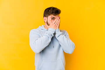 Young caucasian man isolated on yellow background blink through fingers frightened and nervous.