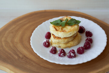 Juicy curd cheesecakes lie on a white saucer with raspberries and mint leaf. Light background