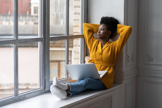 Relaxed African American millennial woman with afro hairstyle wear yellow cardigan, sitting on windowsill, resting, taking break from work on laptop, thinking and looking at window, hands behind head.