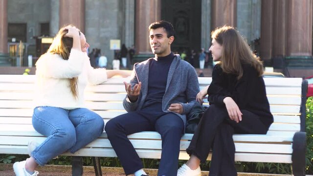 Two young girls and a man friends sitting on a bench in summer park chatting happily having fun. Media. Portrait of two smiling women and a man spend time together and talking.