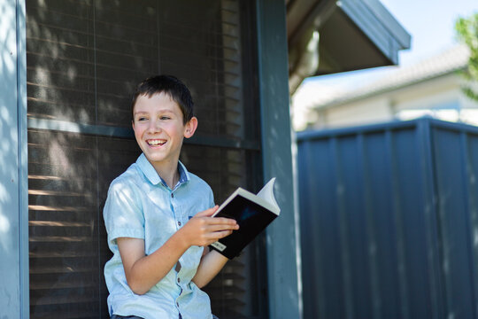 Boy laughs as he reads book outside next to house window