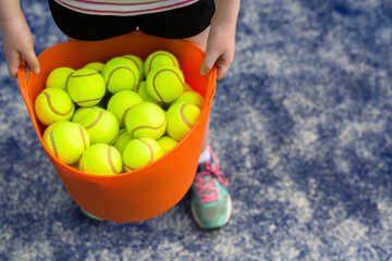 Girl holding an orange basket of tennis balls on a blue tennis court