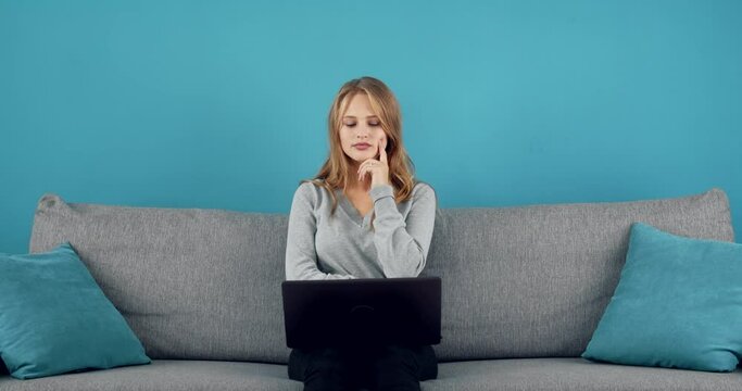 Thoughtful young woman with blond hair sitting on cozy sofa and looking at computer screen. Attractive girl in grey sweater using wireless laptop over blue background.