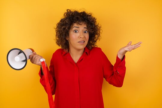 Clueless Young arab woman with curly hair wearing red shirt holding a megaphone over yellow background, shrugs shoulders with hesitation, faces doubtful situation, spreads palms, Hard decision