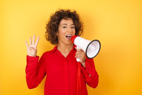 Young arab woman with curly hair wearing red shirt holding a megaphone over yellow background showing and pointing up with fingers number four while smiling confident and happy.