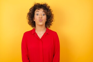 Shot of pleasant looking Young arab woman with curly hair wearing red shirt  on yellow background  pouts lips, looks at camera, has long hair, , has fun with girlfriend,  Human facial expressions