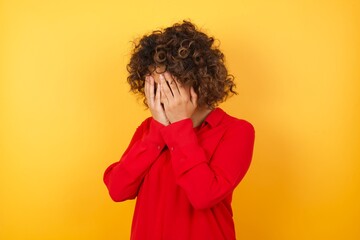 Fototapeta na wymiar Sad Young arab woman with curly hair wearing red shirt on yellow background crying covering her face with her hands.