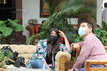 
Latino man and woman with protection mask talking in living room with natural vegetation, new normal covid-19