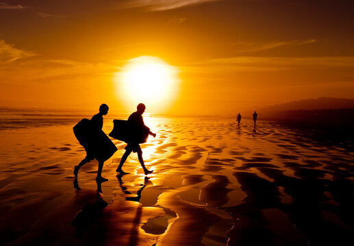 Surfing Sunset. Silhouette of two surfers carrying boogie boards along the sand of a beach.