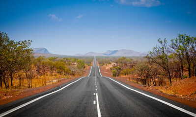 Fototapeta na wymiar The open road in Kimberly, Western Australia. Straight single lane asphalt road stretching into the distance with mountains in the background. Holiday adventure. 