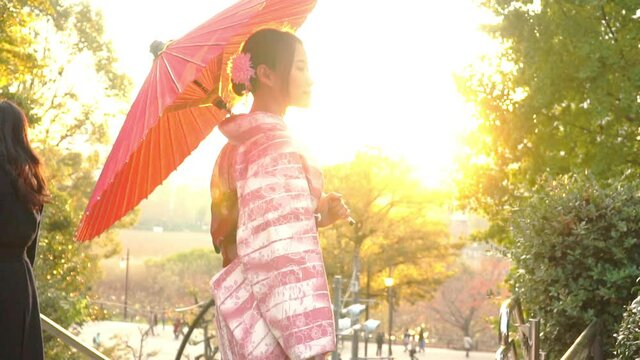 Slow motion of Smiling young beautiful Asian woman tourist in pink traditional japanese kimono dress holding red umbrella relax and enjoy travel vacation in autumn at public park in Tokyo city, Japan