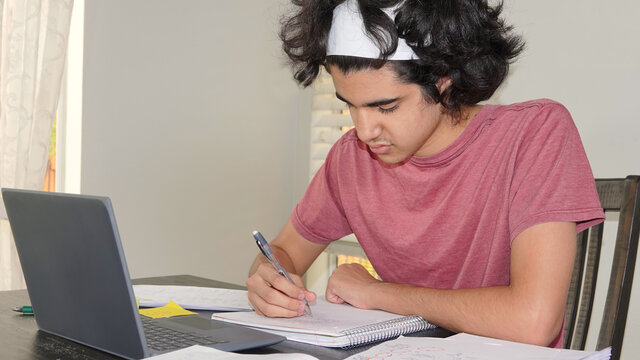 Teen boy of Persian ethnicity doing remote school in times of Covid-19, taking math notes from a virtual lesson on his laptop. 