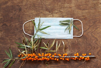 Wooden natural background with a medical protective mask with sea buckthorn branches, with berries. The idea of protecting against the flu with folk natural remedies.