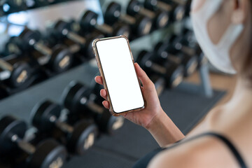 Image of woman wearing face mask and her hand holding mobile phone with white screen mockup at gym.