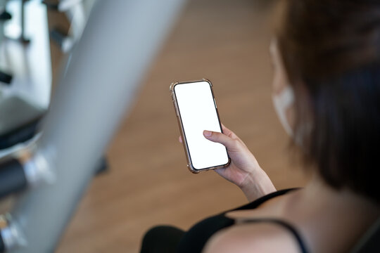 Image of woman wearing face mask and her hand holding mobile phone with white screen mockup at gym.