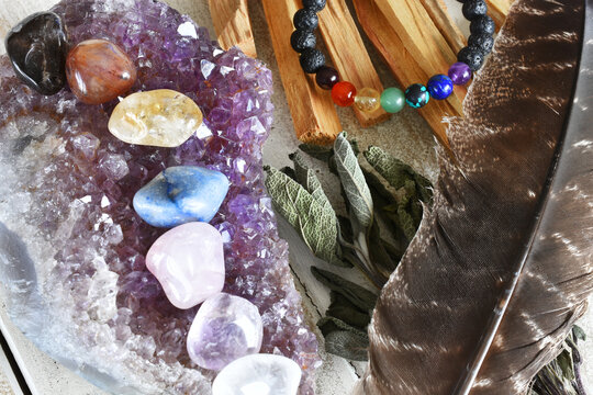 A close up image of chakra crystals charging on an amethyst geode with sage and palo santo smudge sticks. 