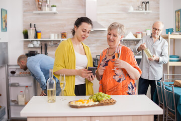 Mother and daughter using smarthphone in kitchen. Senior woman holding glass of wine. Old man looking at bottle.