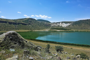The volcanic Lake Nar is surrounded by mountains. Boulders lie on the shore, grass grows. Blue sky. Reflection in calm emerald water. Turkey.