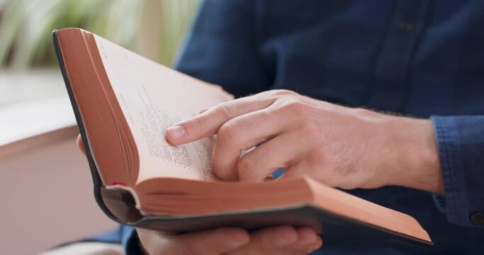 Closeup view of male hands holding book. Young man is reading a book while sitting in white chair, at home. Window and green plants on background.